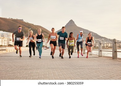 Diverse Group Of Young People Running Along A Seaside Promenade. Fit Young Runners Training Outdoors By The Seaside.