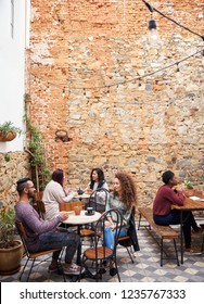 Diverse Group Of Young People Talking Over Coffee While Sitting Together At Tables In The Courtyard Of A Trendy Cafe