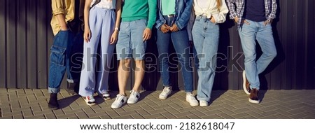 Diverse group of young people wearing jeans. Six men and women in comfortable blue jeans, shorts, and trendy sneakers leaning on street wall. Banner, crop shot, human legs. Casual city fashion concept