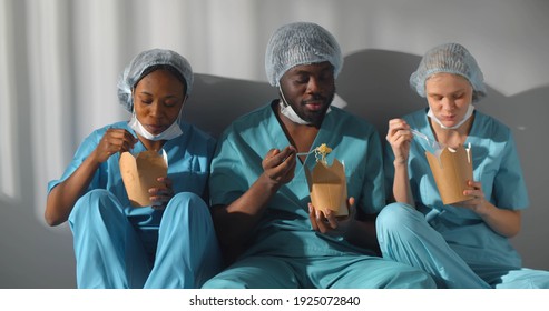 Diverse Healthcare Workers In Scrubs Uniform Talking And Eating Takeaway Food Sitting On Floor In Hospital Corridor. Multiethnic Medical Staff Having Meal During Break