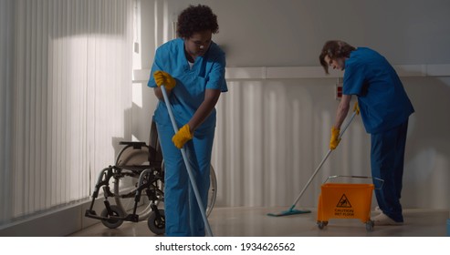 Diverse Janitors Washing Floor In Hospital Room With Empty Wheelchair. Multiethnic Nurse Team Cleaning Floor With Mop And Bucket In Clinic. Healthcare And Hygiene Concept