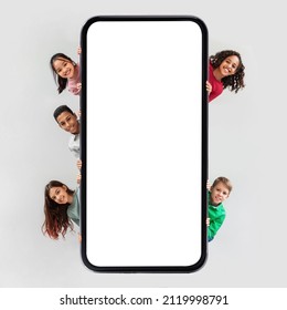 Diverse Kids Posing Near Huge Phone Blank Screen Hiding And Smiling To Camera Advertising Mobile Application Over Gray Studio Background. Children And Gadgets Concept. Square Shot, Mockup