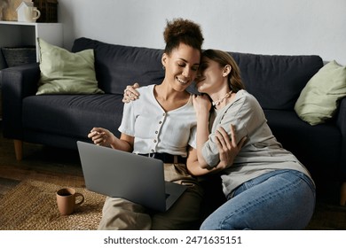 A diverse lesbian couple cuddles together on the floor, smiling and laughing while using a laptop. - Powered by Shutterstock