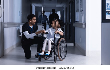Diverse male doctor talking with girl in wheelchair in hospital corridor. Medicine, healthcare, childhood and medical services, unaltered. - Powered by Shutterstock