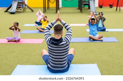 Diverse male teacher and elementary schoolchildren practicing yoga in schoolyard. Education, inclusivity, elementary school and learning concept. - Powered by Shutterstock