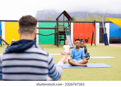 Diverse male teacher and elementary schoolchildren practicing meditation in schoolyard. Education, inclusivity, elementary school and learning concept. - Powered by Shutterstock