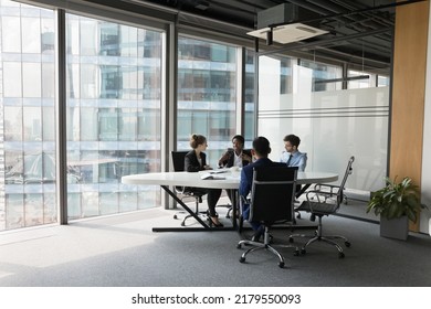 Diverse millennial business team talking in meeting room, negotiating on project at table at glass wall panoramic window, discussing deal in open space, modern office interior. Wide shot
