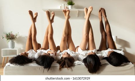 Diverse millennial girls lie upside down show legs celebrate bridal shower at home together, multiracial female friends in bathrobes have fun at spa pajama hen party in hotel, depilation concept