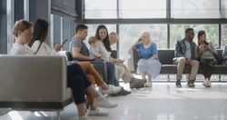 Diverse Multicultural People Sit On Couches In Clinic Lobby Area, Wait For Appointment With Doctor Or Medical Test Results. Waiting Area In Medical Center With Modern Design. Healthcare System.