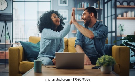 Diverse Multiethnic Couple are Sitting on a Couch Sofa in Stylish Living Room and Choosing Items to Buy Online with Laptop Computer, Give High Five. Friends or Colleagues are Discuss Work Projects.