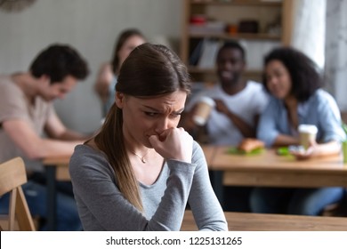 Diverse multi-ethnic friends sitting together in cafe talking having fun, focus on frustrated shy girl sitting separately by others teenagers feels unhappy because peers not accept her she is outcast