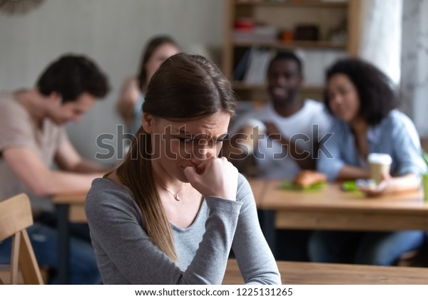 Diverse multi-ethnic friends sitting together in\
cafe talking having fun, focus on frustrated shy girl sitting\
separately by others teenagers feels unhappy because peers not\
accept her she is\
outcast