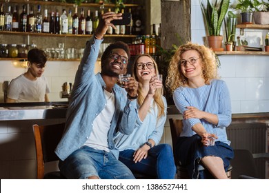 Diverse Multiethnic Group Of Friends Taking Selfie On Smart Phone. Young Black Men And Two Women Sitting On Stools Near Counter And Taking A Self Portrait On Cell Phone.