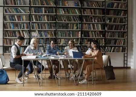 Diverse multiethnic group of young college students cooperating on graduation project in library, discussing research study, sitting at table with books, sharing learning tasks. Wide shot