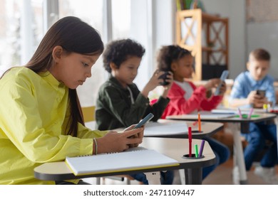 Diverse multiethnic kids students having break and using smartphones, sitting at desks in classroom, children playing games and checking social media at school - Powered by Shutterstock