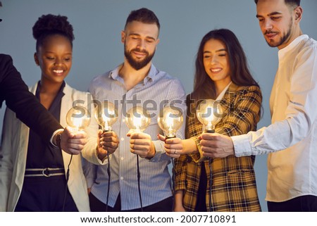 Diverse multinational group of smiling people holding illuminated lightbulb joining together brainstorming and sharing creative idea on startup project studio shot on grey background