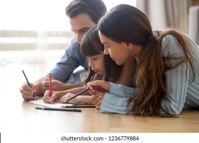 Diverse Multiracial Family Married Couple With Little Adorable Daughter Lying On Wooden Warm Floor At Modern Home Drawing Together On Paper Using Colored Pencils. Child Development And Hobby Concept