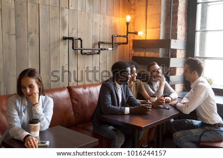 Diverse multiracial people hanging together in coffeehouse ignoring sad young girl sitting alone at cafe table, upset social outcast loner suffers from unfair attitude or discrimination among friends