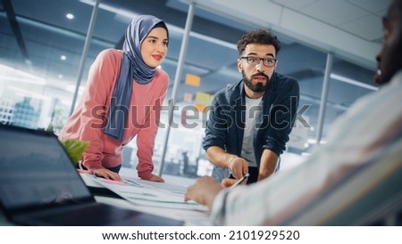 Diverse Office Big Table Meeting: Muslim Businesswoman Wearing Hijab and Hispanic Male Entrepreneur Work with Documents, Talk, Brainstorm. Professionals in e-Commerce Project. Dutch Angle