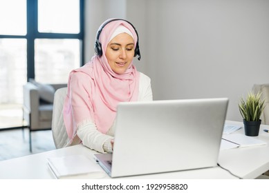 Diverse office employees concept. A focused young muslim woman wearing hijab and smart casual wear using headset and the laptop in contemporary office space