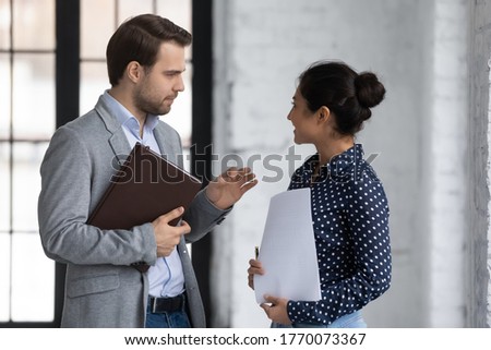 Diverse office workers colleagues met standing in office and talking, friendly Indian woman communicates with Caucasian man discussing project ideas, 2 executives having pleasant conversation concept