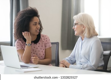 Diverse old and young female colleagues talking at work, african and caucasian business women sitting together in office having friendly conversation, mentor intern discussing planning shared project