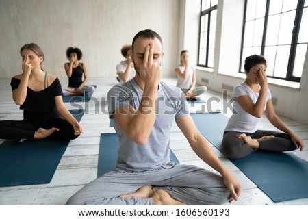 Diverse people doing Alternate Nostril Breathing exercise, practicing yoga at group lesson, nadi shodhana pranayama, sitting in Lotus pose on mats, working out in modern yoga studio, center