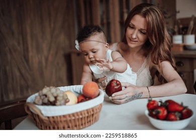 Diverse people portrait of mother with swarthy infant takes fruits sitting at table. Multi ethnic family together enjoy healthy breakfast vegan food in positive emotion and motherly care at home - Shutterstock ID 2259518865