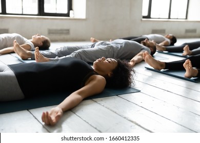 Diverse People Resting After Work Out Lying On Carpets Performing Shavasana Or Corpse Dead Position Focus On African Female, Final Pose Of Yoga Class Deep Restoration, Body And Mind Relaxation Concept