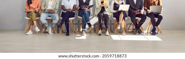 Diverse people in smart casual clothes holding\
paper documents, CVs, resumes, laptops and cellphones waiting in\
line for job interviews or business appointments in recruitment\
agency, legs low\
section