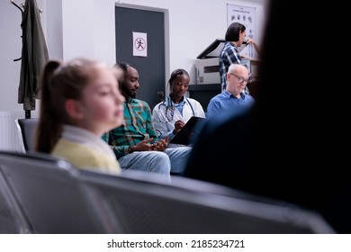 Diverse People Waiting In Private Clinic While Hospital Medic Is Asking Medical Questions In Waiting Room. Selective Focus On African American Doctor With Stethoscope Completing Form For Patients.