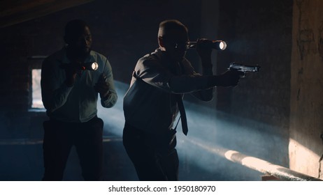 Diverse Policemen With Flashlight And Gun Chasing Criminal In Dark Attic. Portrait Of Multiethnic Detectives Searching Evidence And Catching Murderer At Crime Scene