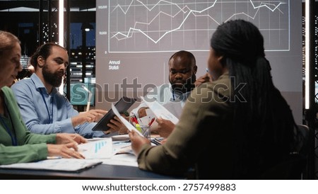 Diverse professionals group evaluating files and performance indicators, using projections and diagrams to align the company objectives with investors expectations for company progress. Camera A.
