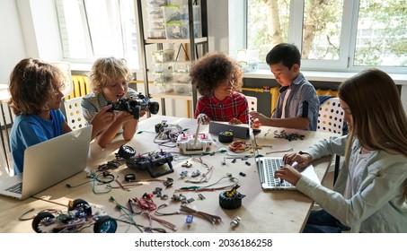Diverse school children students group building robotic cars using computers. Happy multicultural kids learning programming robot vehicles sitting at table at STEM education coding engineering class.