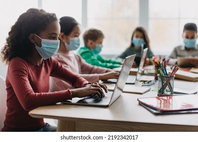 Diverse School Kids Using Laptop Computers Learning Online And Wearing Face Masks For Covid-19 Protection Sitting In Modern Classroom Indoor. Selective Focus