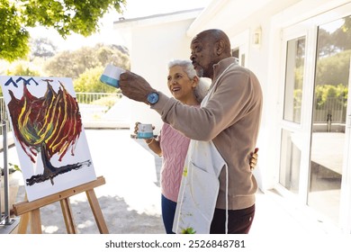 Diverse senior couple drinking coffee, talking and embracing next to painting on sunny terrace. Lifestyle, retirement, senior lifestyle, nature, creativity, togetherness and domestic life, unaltered. - Powered by Shutterstock