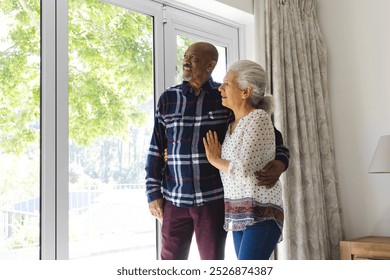 Diverse senior couple embracing and looking out window at sunny home. Lifestyle, retirement, senior lifestyle, togetherness and domestic life, unaltered. - Powered by Shutterstock