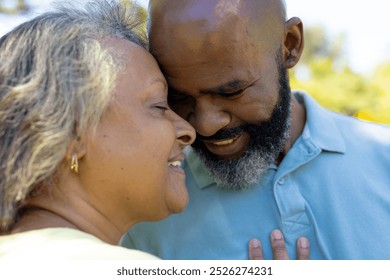 A diverse senior couple embracing outdoors, sharing affection. Both showing happiness, smiling in sunny park with a green background, unaltered - Powered by Shutterstock