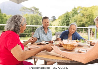 Diverse senior female friends enjoying meal outdoors, saying grace and praying. Wearing casual clothing, sharing conversation, showing signs of aging, with green background, unaltered - Powered by Shutterstock