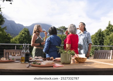 Diverse senior female friends enjoying food outdoors, one tasting snack. Wearing casual clothing, laughing and chatting with blue background, unaltered - Powered by Shutterstock