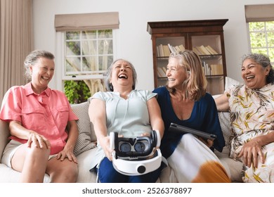 Diverse senior female friends laughing together on couch at home. Holding a virtual reality headset and a tablet, enjoying new technology and sharing joyful moments, unaltered - Powered by Shutterstock