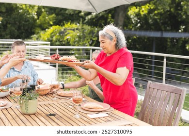 Diverse senior female friends sharing meal outdoors. Biracial woman in red serving, others with light hair and skin, all in casual wear, enjoying food together, unaltered - Powered by Shutterstock