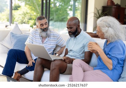 Diverse senior friends gathering around laptop at home. African American man and Caucasian couple discussing content, sitting on a blue background couch, unaltered - Powered by Shutterstock