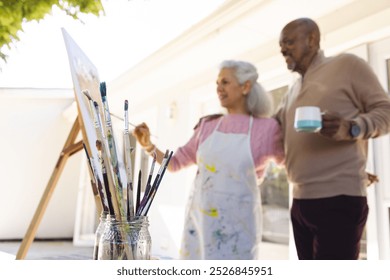 Diverse senior woman painting on canvas and senior man embracing her on sunny terrace. Lifestyle, retirement, senior lifestyle, nature, creativity, togetherness and domestic life, unaltered. - Powered by Shutterstock