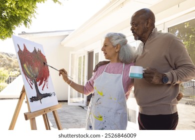 Diverse senior woman painting on canvas and senior man embracing her on sunny terrace. Lifestyle, retirement, senior lifestyle, nature, creativity, togetherness and domestic life, unaltered. - Powered by Shutterstock