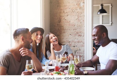 Diverse Small Group Of Four Happy Friends Having Wine And Dinner Together In Restaurant With Brick Wall And Bright Large Window In Background