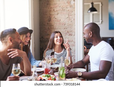 Diverse Small Group Of Four Happy Friends Having Wine And Dinner Together In Restaurant With Brick Wall And Bright Large Window In Background