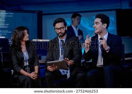 A diverse software engineering team in a SOC. An Indian developer holding a tablet discusses AI algorithm optimization and cybersecurity protocols. His team, including women and men, are in a briefing.
