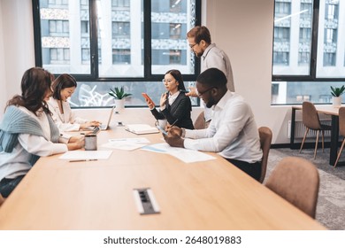 Diverse startup team working together around table with laptops, smartphones, documents and coffee cups in modern office, collaborating on tech project in creative coworking space