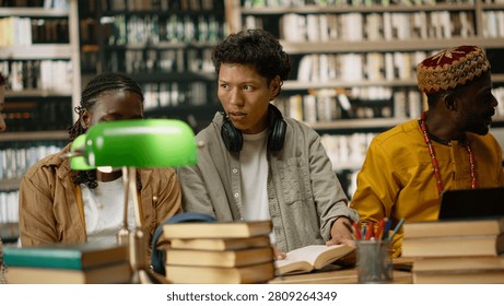 Diverse students discuss classwork during group study around library table with open books, textbooks and green lamps, collaborative academic study, peers collaboration.